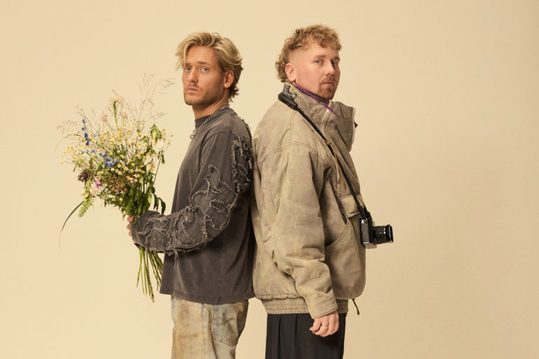 Bandet Norlie & KKV står rygg mot rygg. En håller i blombukett och en annan har en kamera runt halsen.