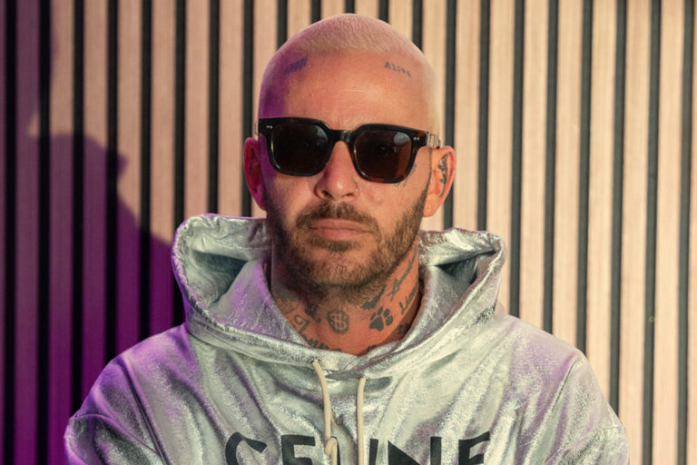 Artisten Joakim Lundell står med armarna i kors, mörka glasögon och grå huvtröja.